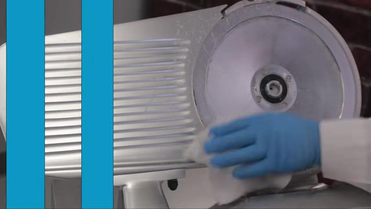 Operator wiping the grooved body and blade area of the slicer with a cloth in the food preparation area.