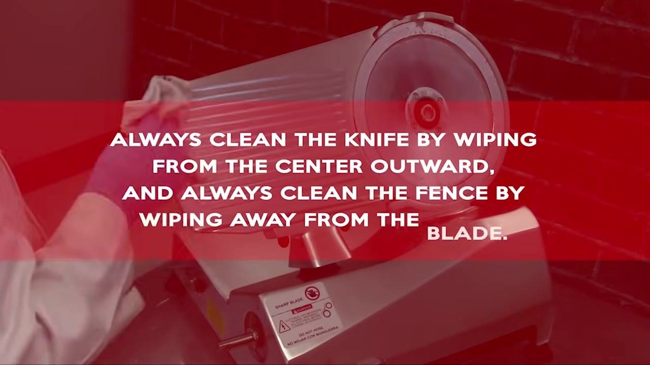 Operator cleaning the meat slicer blade with a cloth; on-screen text reads "ALWAYS CLEAN THE KNIFE BY WIPING FROM THE CENTER OUTWARD, AND ALWAYS CLEAN."