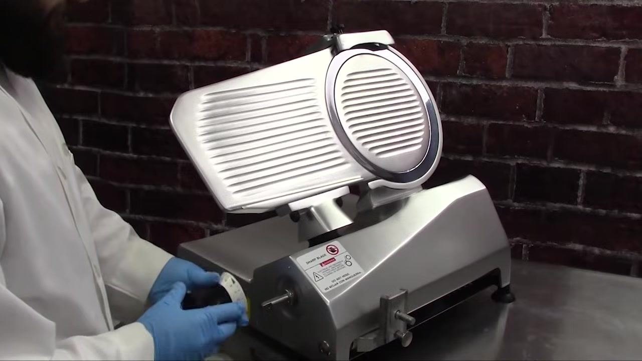 Operator holding the slicer sharpener above the slicer, preparing to reinstall it into its storage position; the slicer is fully assembled except for the sharpener.