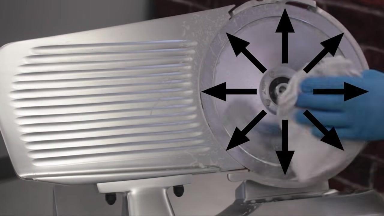 Operator cleaning the slicer blade with a cloth, following outward arrows indicating a circular motion from the center.