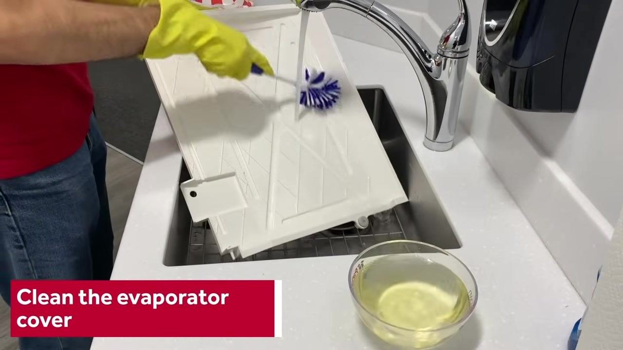 Person wearing yellow gloves scrubbing the evaporator cover in a sink with a nylon brush, cleaning solution nearby, with the on-screen instruction "Clean the evaporator cover"