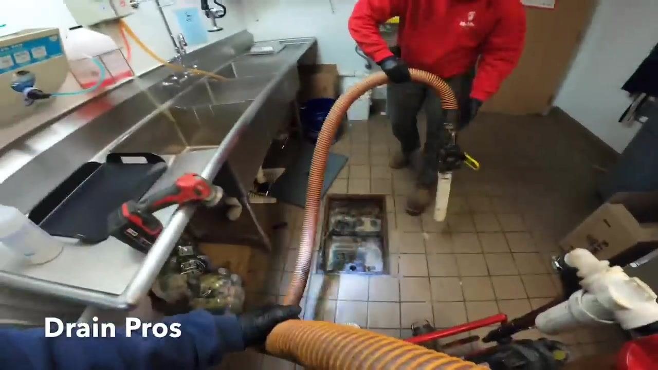 Technicians using a vacuum hose to clean out a grease trap in a commercial kitchen, with gloves, boots, a stainless steel sink, and the open grease trap visible.