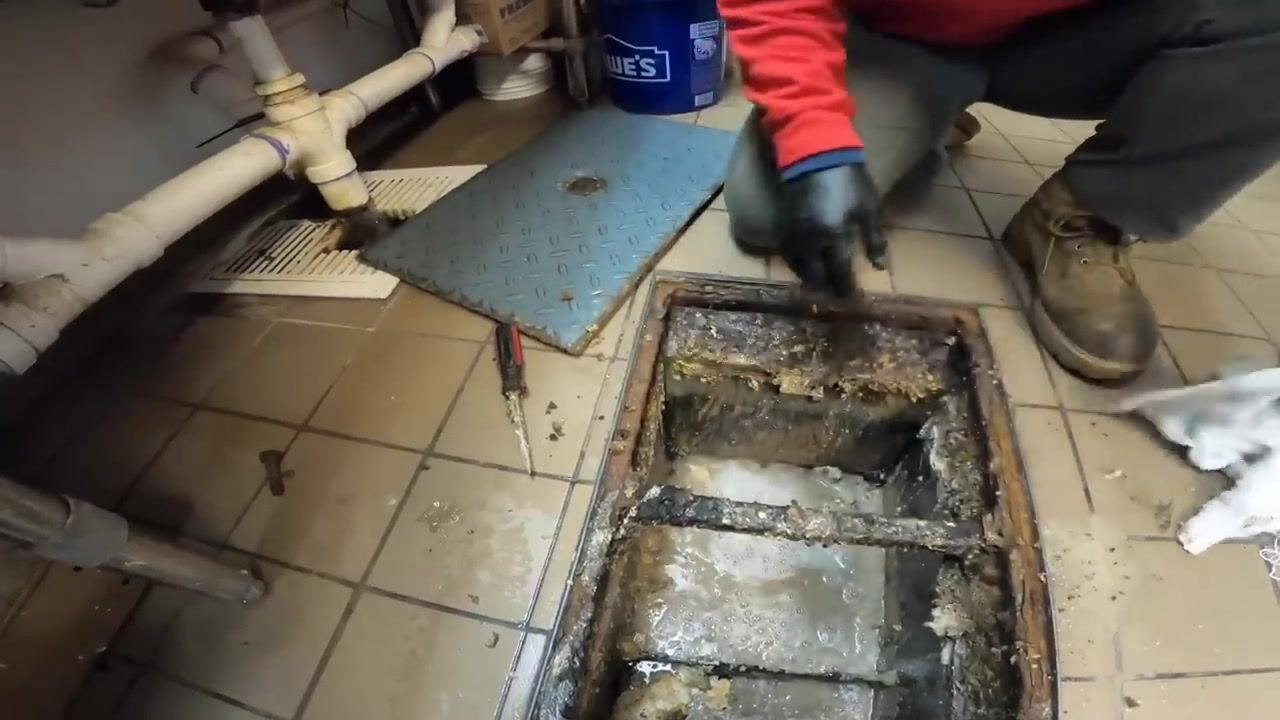 Technician kneeling beside an open grease trap, reinstalling the baffle. The trap is visibly dirty, with tools and cleaning supplies nearby.
