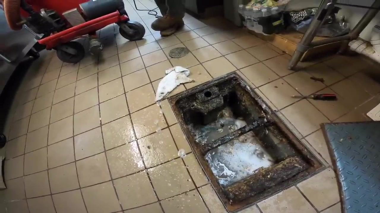 Open grease trap with water and foam inside, surrounded by cleaning equipment and technicians, with the area wet from recent cleaning and testing.