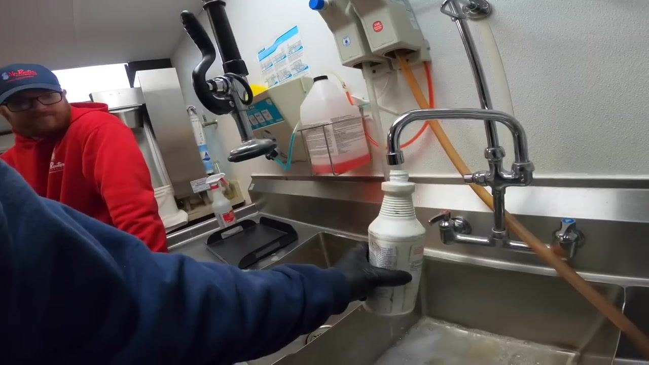 Gloved hand pouring cleaning solution into a stainless steel sink while water runs from the faucet, with various cleaning supplies and dispensers visible.