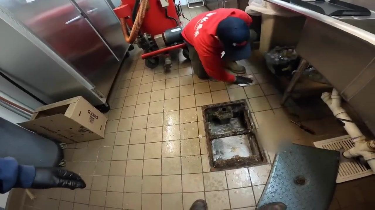 Technician in red uniform kneeling and cleaning the area around an open grease trap on a tiled floor, with a vacuum, cleaning equipment, and a cardboard box nearby.