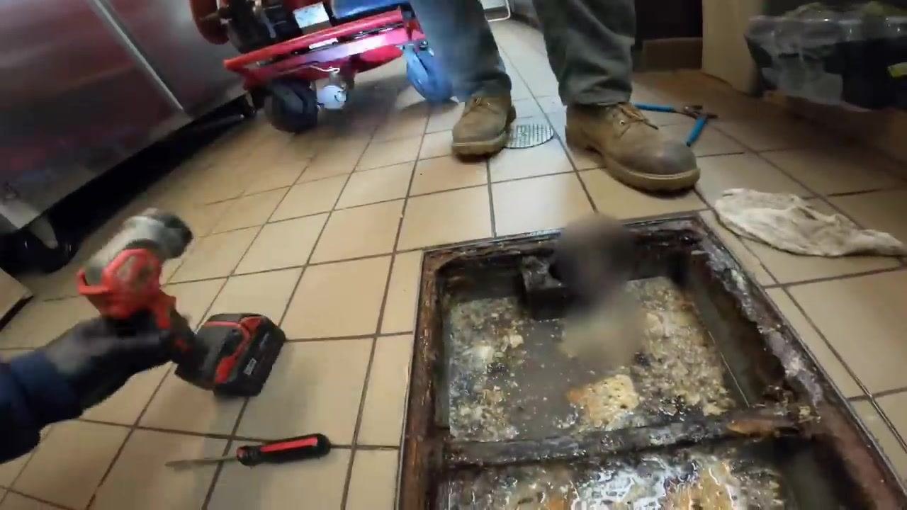 Technician's gloved hand placing a bolt into the metal cover of the grease trap, with the cover partially secured and cleaning equipment visible in the background.