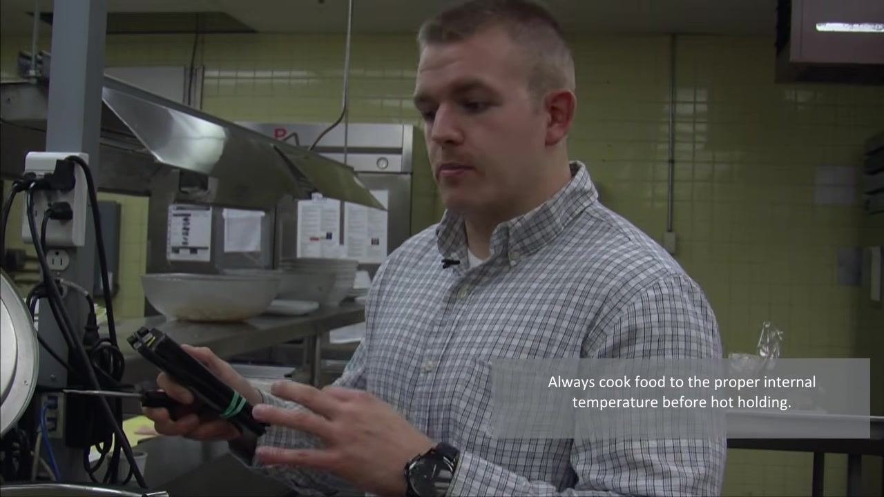 A person holding a thermometer and emphasizing proper cooking and holding temperatures, with on-screen text reading "Always cook food to the proper internal temperature before hot holding."