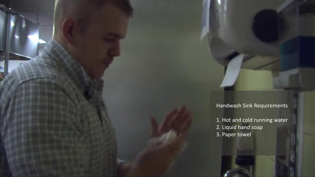 A person drying their hands with paper towel at the handwashing station, with the requirements listed on screen