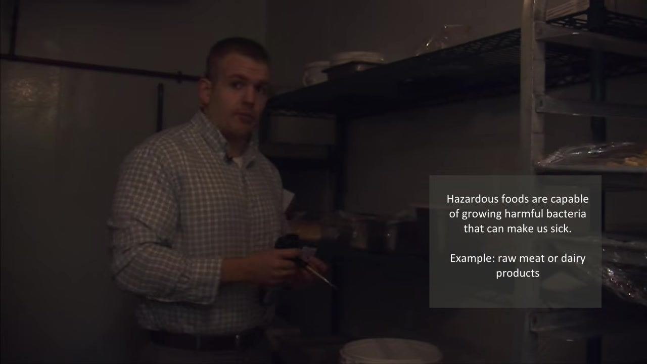 A person holding a thermometer and an alcohol swab inside a walk-in refrigerator, with a reminder about hazardous foods visible on screen