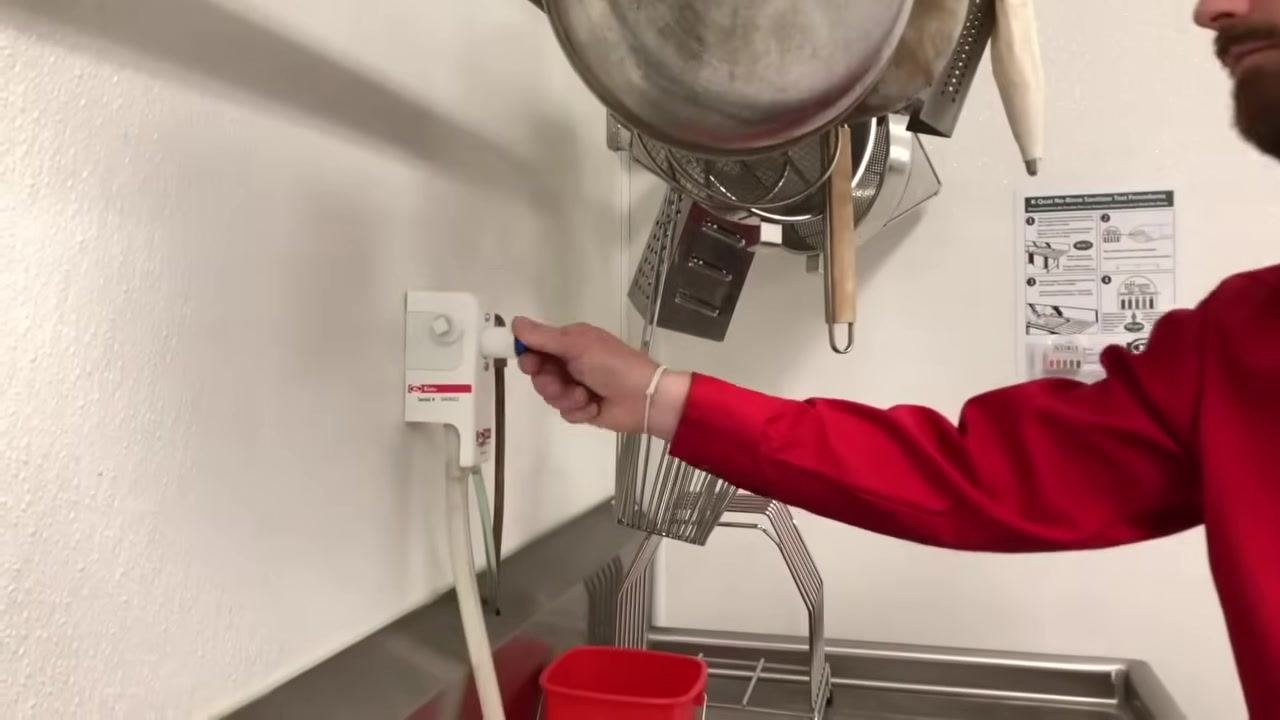 Staff member turning the blue-handled knob on the wall-mounted chemical dispenser above the sink, with utensils and pots hanging nearby