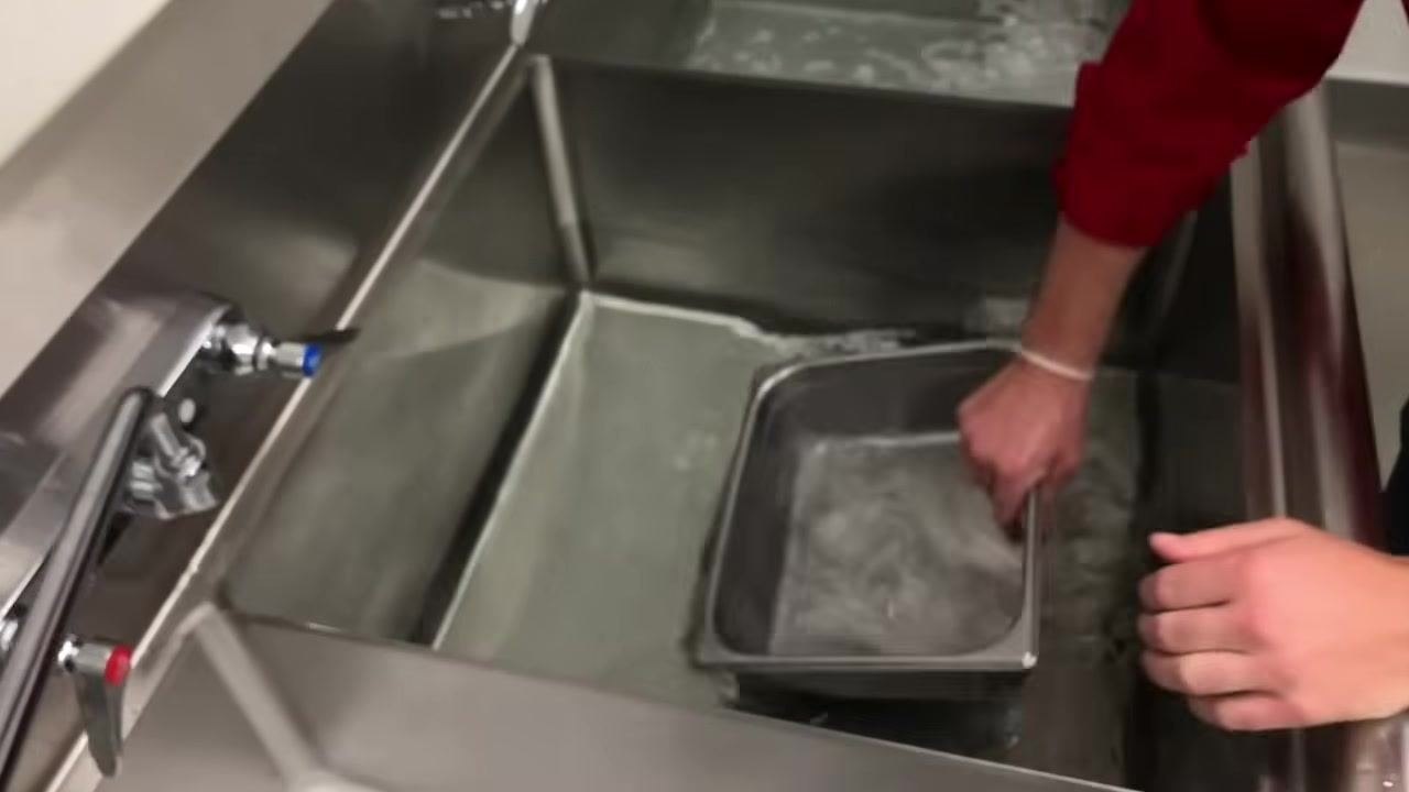 Staff member rinsing a metal container in the rinse compartment, agitating it by hand in clear water