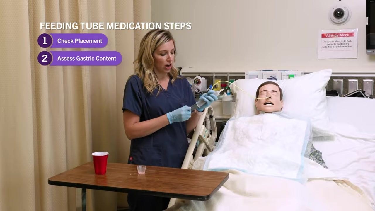 Nurse holding a syringe without a plunger, preparing to attach it to the feeding tube, with on-screen steps 1 and 2 displayed.