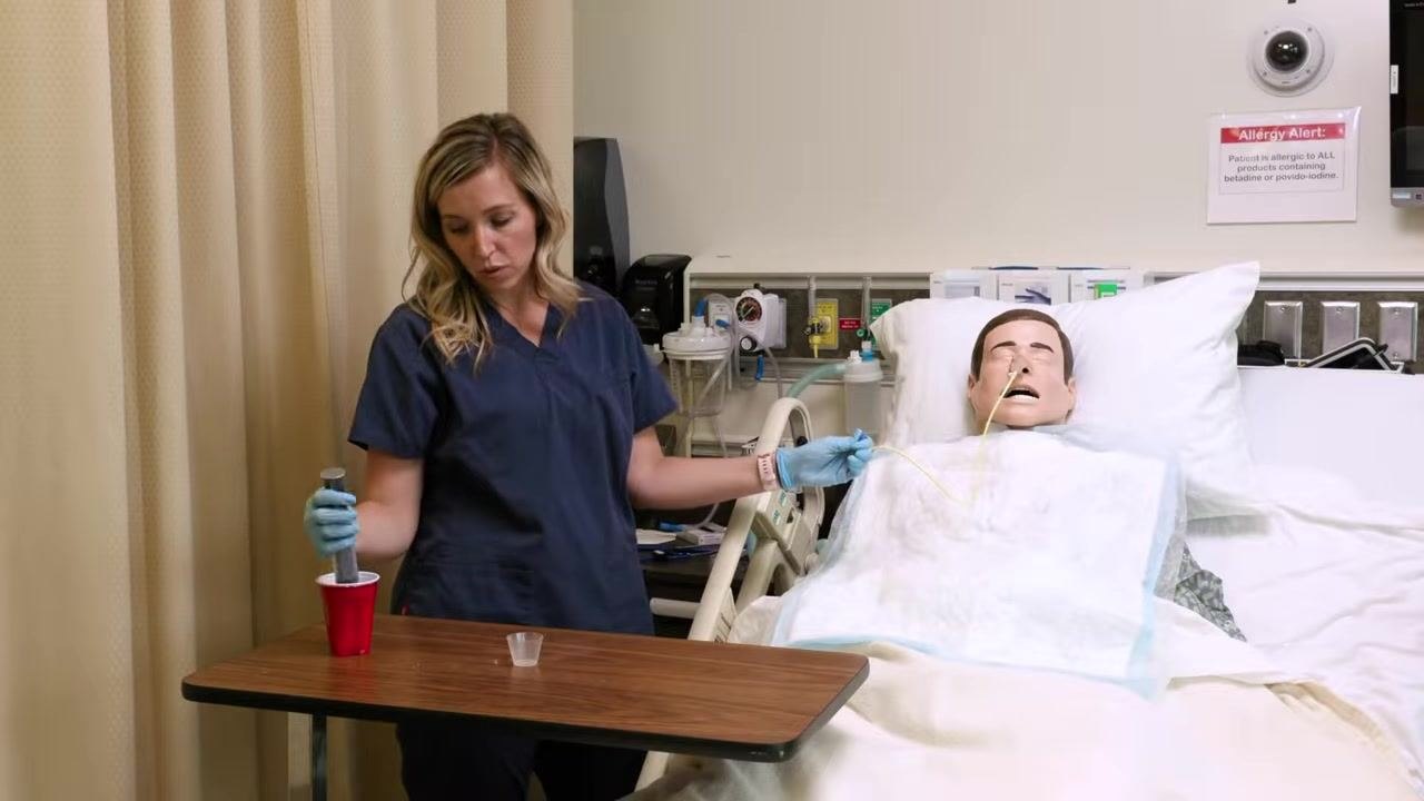 Nurse in blue scrubs holding a syringe with plunger over a red cup, preparing to push water into a feeding tube connected to a mannequin. A medicine cup and supplies are on the table.