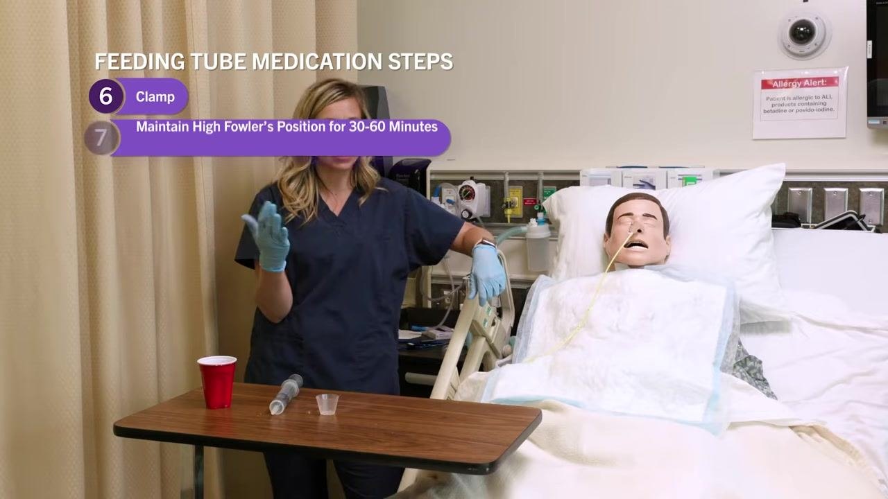 Nurse at the bedside holding the feeding tube and syringe, explaining the next steps. Red cup, medicine cup, and syringe are on the table.