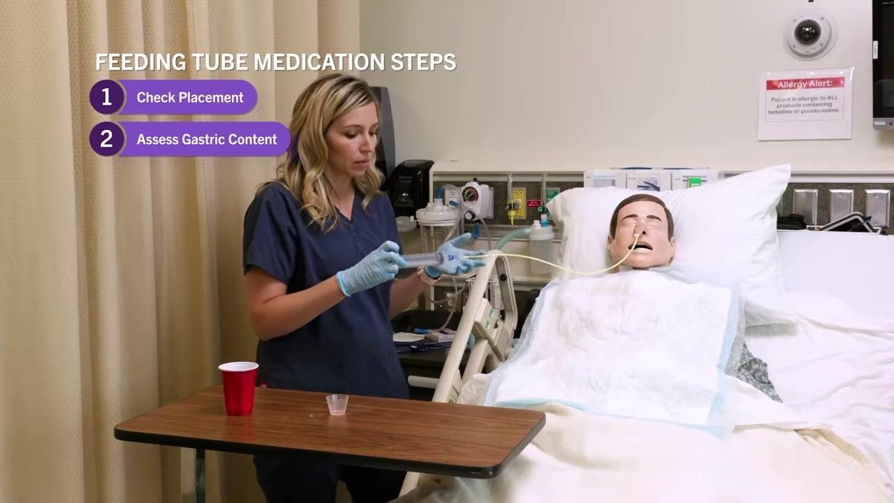 Nurse in navy scrubs holding the NG tube and 60 mL syringe beside a patient mannequin, with on-screen text "Feeding Tube Medication Steps: 1. Check Placement, 2. Assess Gastric Content." Supplies visible on the overbed table.