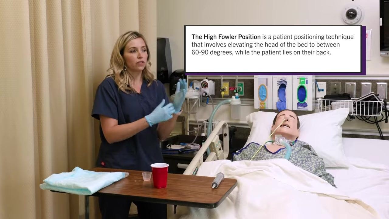 Nurse with gloves and an absorbent pad placed on the patient's chest, medication cup and water cup on the table. Patient mannequin with NG tube is in the bed.