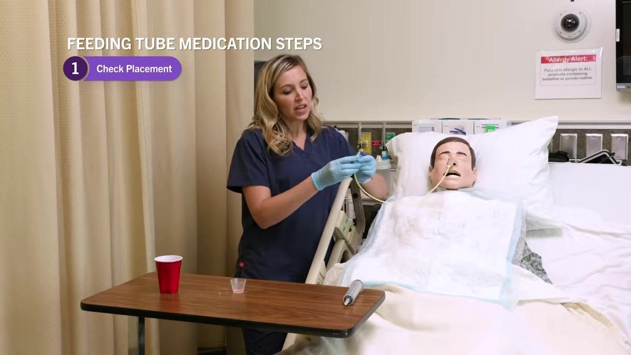 Nurse in navy scrubs holding the NG tube and demonstrating removal of the cap, with on-screen text "Feeding Tube Medication Steps: 1. Check Placement." Patient mannequin is in the bed with supplies nearby.