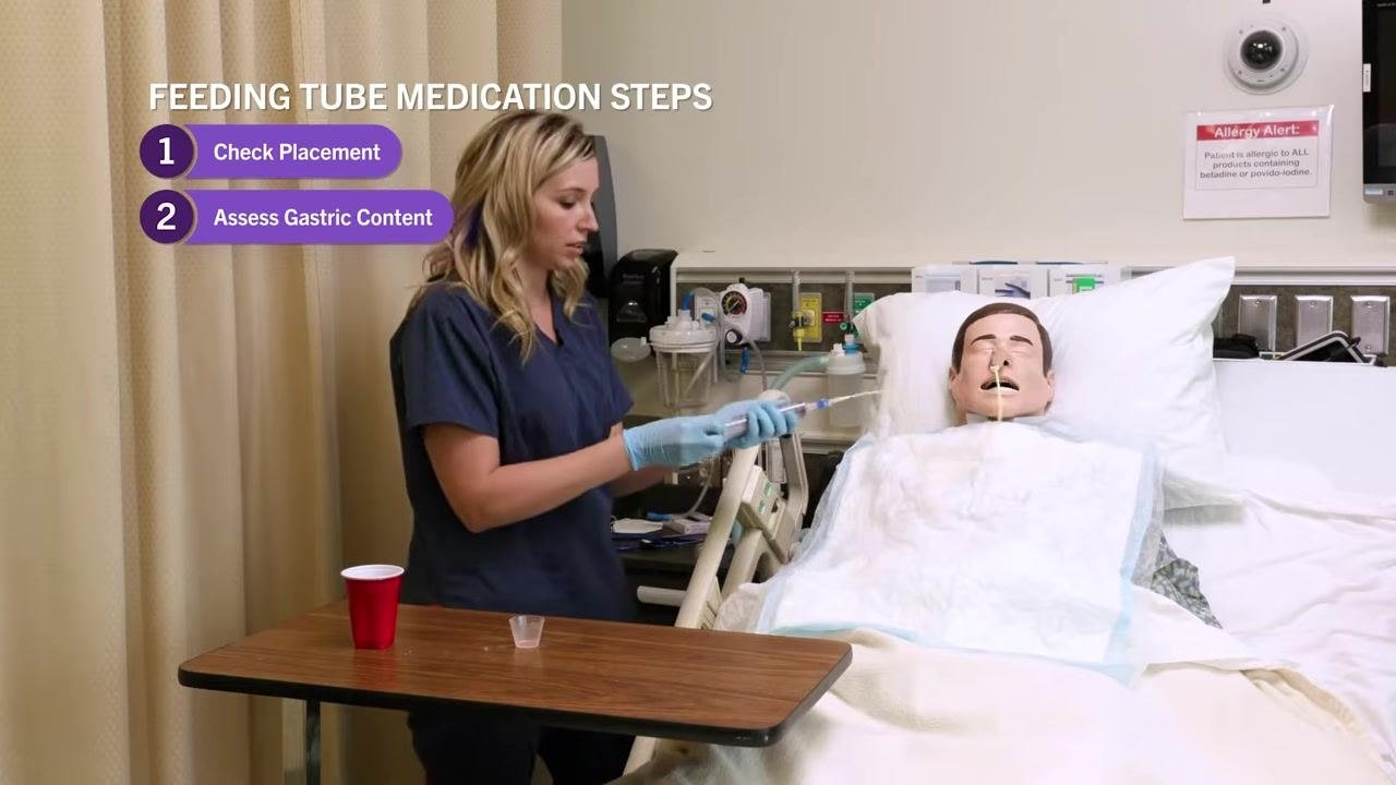 Nurse returning the aspirate to the stomach using the syringe and NG tube, with on-screen text "Feeding Tube Medication Steps: 1. Check Placement, 2. Assess Gastric Content."