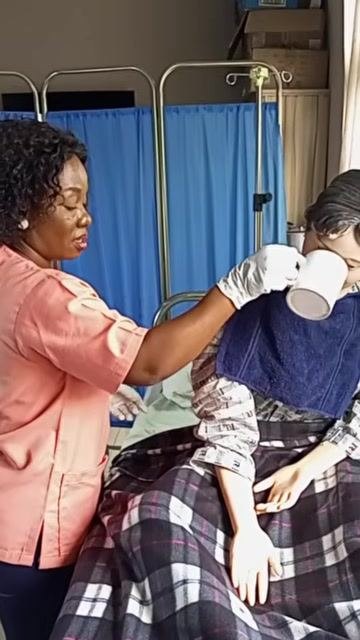 A healthcare worker holding a cup to the patient's mouth so they can rinse, with the patient upright and a towel placed around their neck.