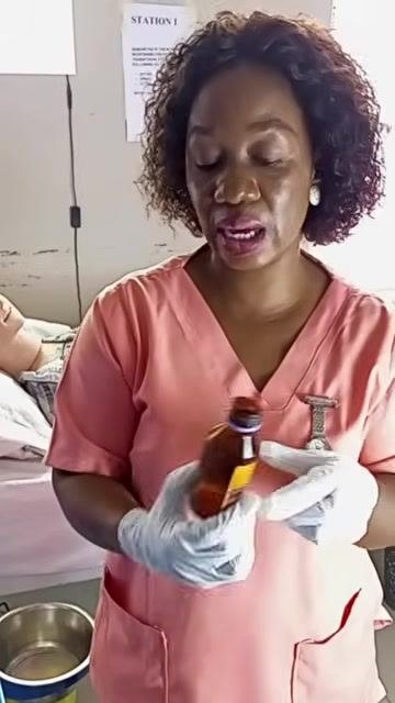 A healthcare worker holding a brown medication bottle at eye level to inspect the label and liquid level, with the patient and a "STATION 1" sign visible in the room.