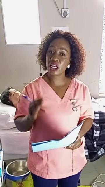 A healthcare worker standing beside the patient's bed, holding a blue chart and gesturing while explaining the medication administration record, with the tray, cups, and water bottle visible nearby.