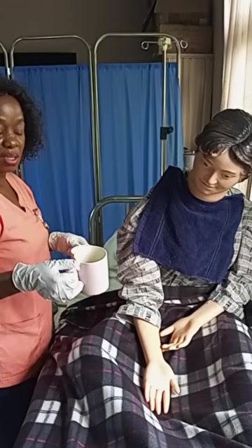 A healthcare worker standing beside the upright patient, holding an empty cup and saucer and preparing to clear away used items while observing the patient for any reaction.