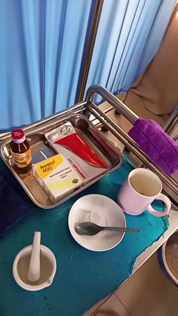 A medical trolley set up with metal bowls, a cup, a mortar and pestle, medication sachets, a bottle, a spoon, and towels, positioned beside a hospital bed with privacy curtains in the background.