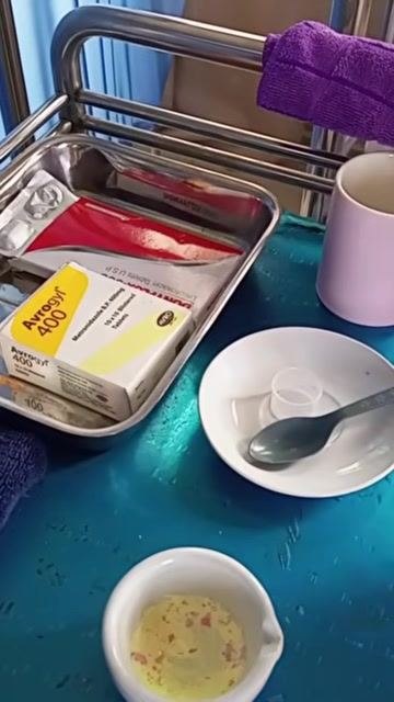 A close-up of medication supplies on the trolley, including syrup, boxed tablets, a mortar and pestle, a bowl with a spoon, and a cup, with gloved hands actively preparing the crushed medication.