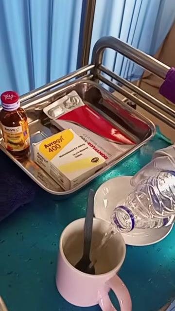 A gloved hand pouring water from a bottle into a cup containing crushed medication, with a second empty cup and saucer prepared for rinsing.