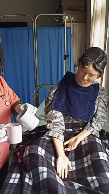 A healthcare worker holding a cup and saucer with the prepared medication mixture, standing beside a patient who is seated upright with a protective towel over their chest.