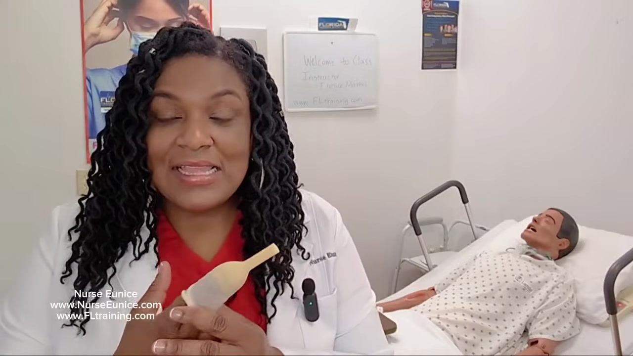 Nurse explaining the procedure and showing the condom catheter, with a mannequin patient in a hospital bed and a whiteboard in the background reading "Welcome to Class, Instructor Eunice Mathis, www.FLtraining.com"