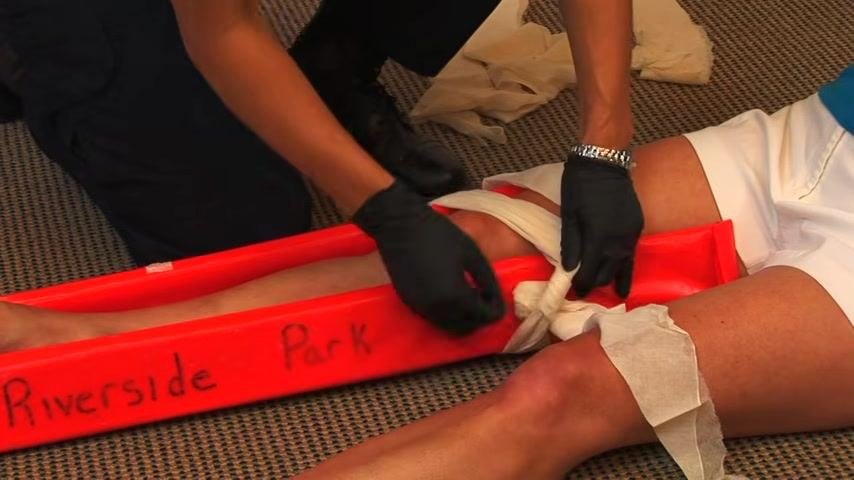 A responder ties a cravat above the fracture site, securing the splint in place. Multiple cravats and the rigid splint are visible.