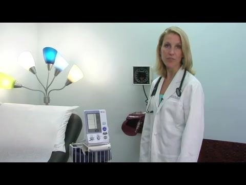 A doctor stands next to a patient bed, with an electronic blood pressure monitor on a cart and a manual wall-mounted aneroid sphygmomanometer nearby. The electronic device has a digital display and labeled buttons in a clinical exam room.