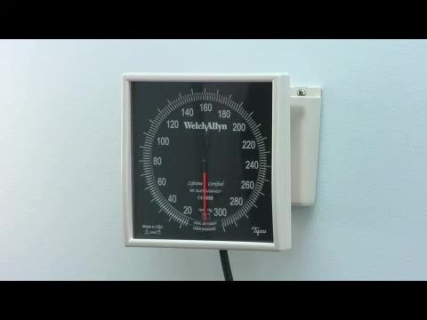 Close-up of a wall-mounted aneroid sphygmomanometer labeled "Welch Allyn" with a black dial and red needle, showing measurement markings from 0 to 300 mmHg.