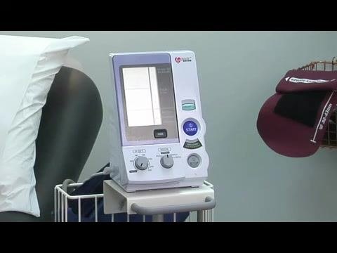 Close-up of an electronic blood pressure monitor on a cart, showing labeled buttons (START, STOP), dials, and a digital display, positioned next to a patient bed in a clinical exam room.