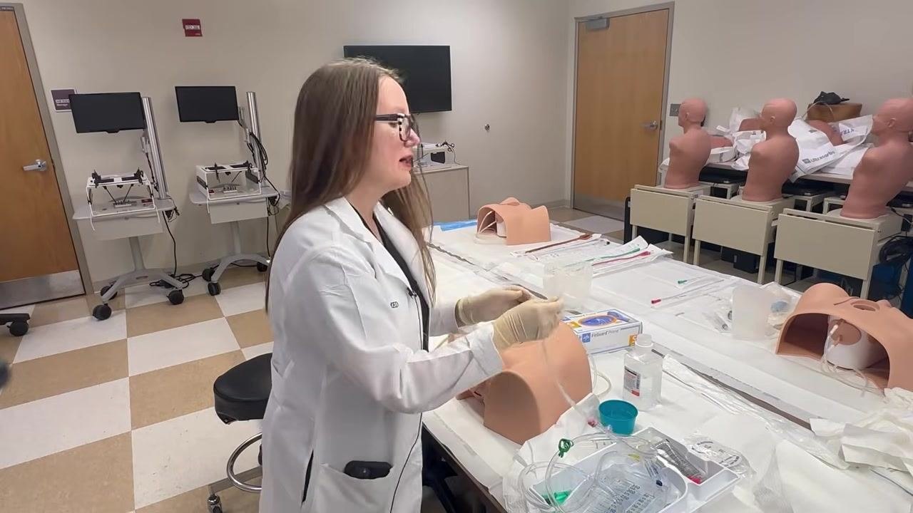 Close-up of hands inflating the balloon of a suprapubic catheter on a training model, with an anatomical diagram inset showing catheter placement in the bladder