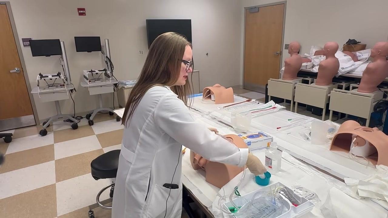 Filling a syringe with saline from a small blue sterile container while holding the catheter in place on a training model