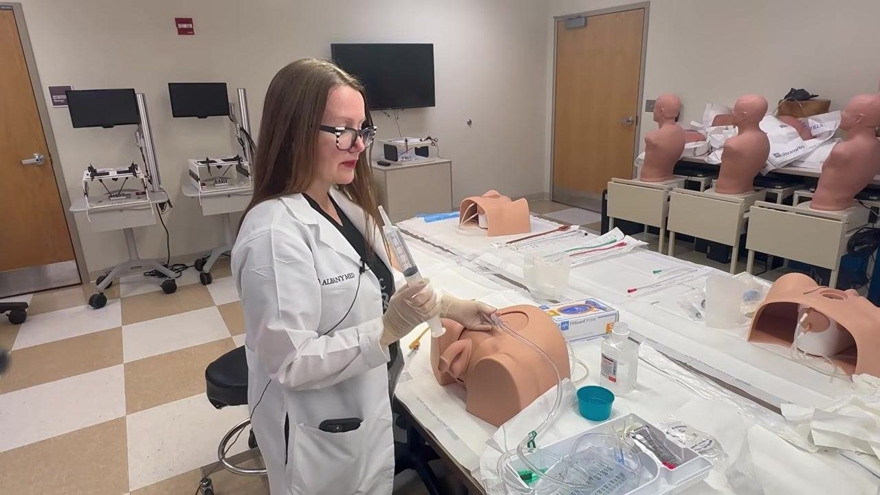 Explaining urine culture collection while holding the catheter and tubing, with supplies visible on the table