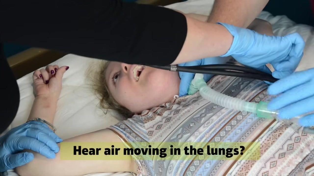 A caregiver uses a stethoscope to listen to the patient's chest while another stabilizes the trach tube. On-screen text: "Hear air moving in the lungs?"