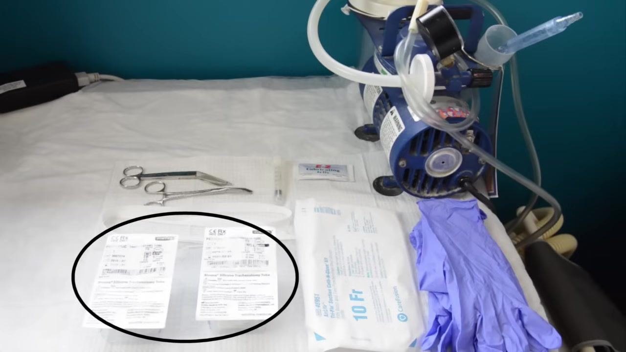 All required supplies for a trach tube change laid out on a clean surface: two trach tube packages, non-sterile gloves, a suction machine, a catheter, lubricating jelly, scissors, tweezers, and a syringe.