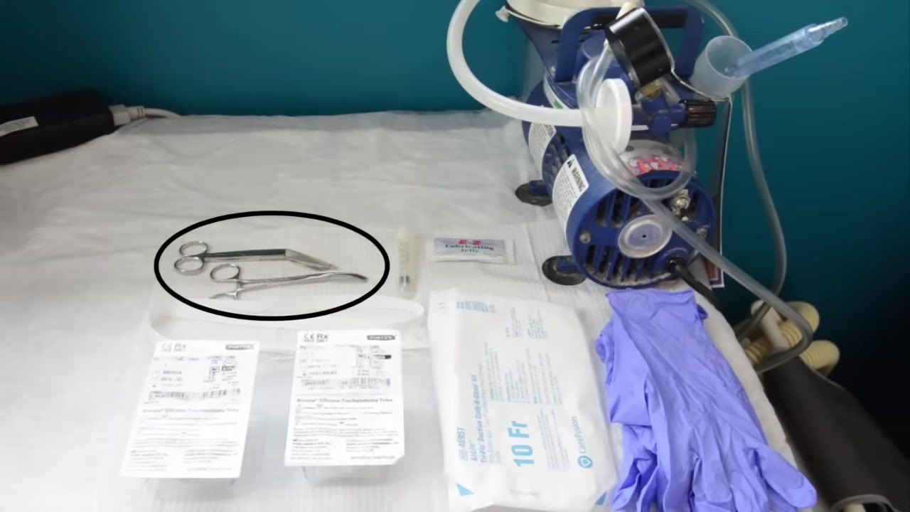 A gloved caregiver applying lubricating jelly to the trach tube, with all supplies visible on the work surface.