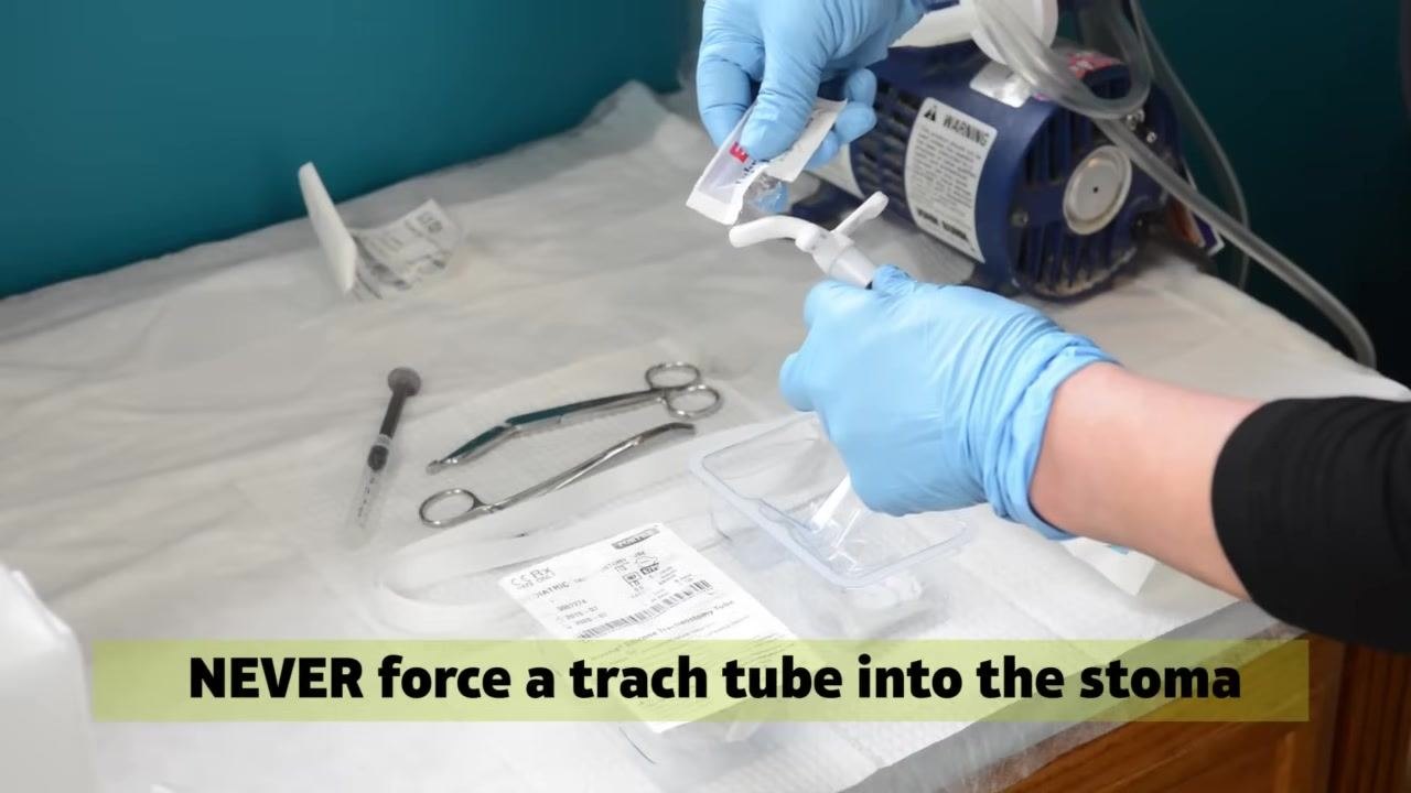 Two caregivers wearing blue gloves: one attaches a Velcro trach tie around the patient's neck, securing it to the trach tube. On-screen text: "Velcro Trach Tie."