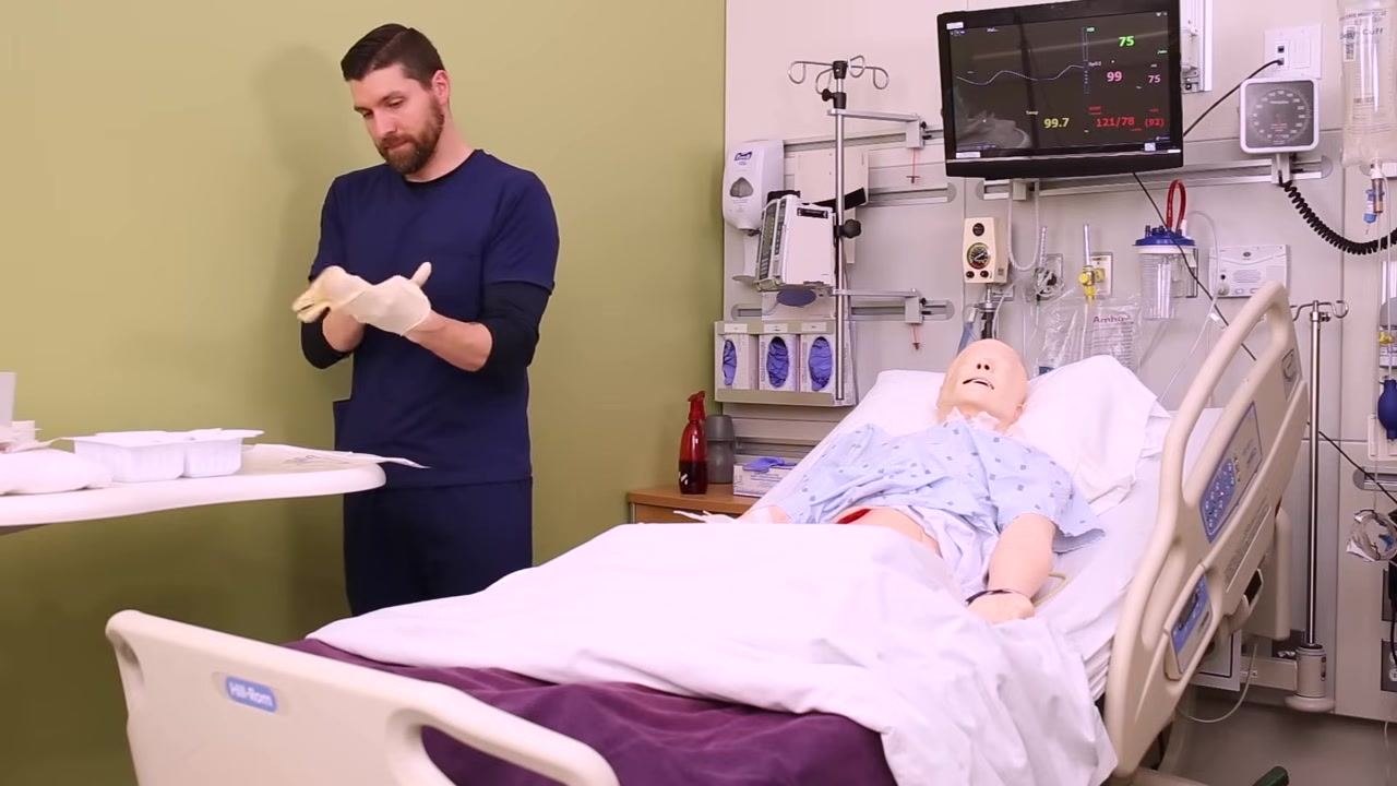 Nurse putting on sterile gloves at the bedside with wound care supplies laid out on a table, patient mannequin in bed, and monitoring equipment visible