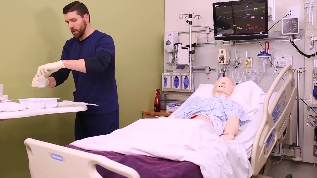 Nurse pouring sterile saline onto gauze pads on a sterile field, with a patient mannequin in a hospital bed and monitoring equipment visible