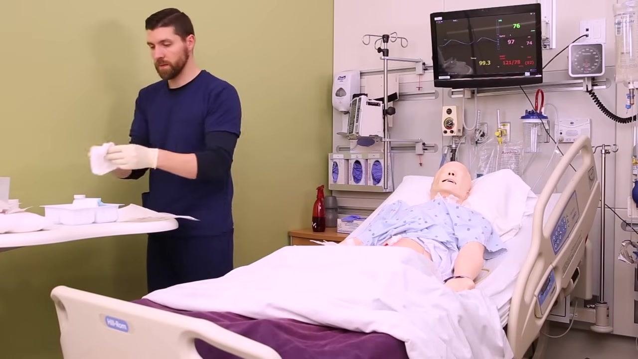 Nurse preparing dry gauze at a bedside table, with a patient mannequin in a hospital bed and monitoring equipment visible