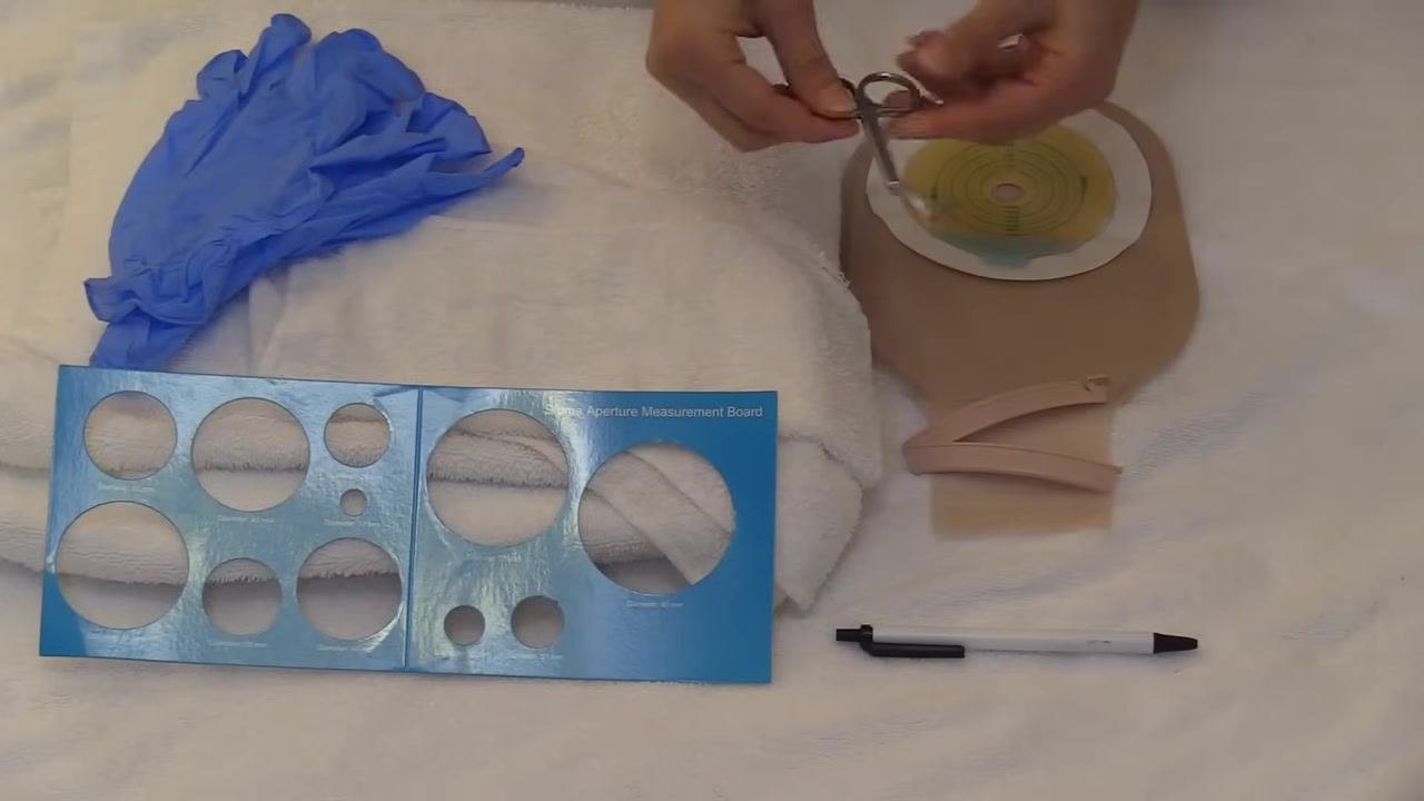 Close-up of supplies including gloves, measurement board, towel, pouching system with wafer, clip, pen, and ostomy scissors held above the towel