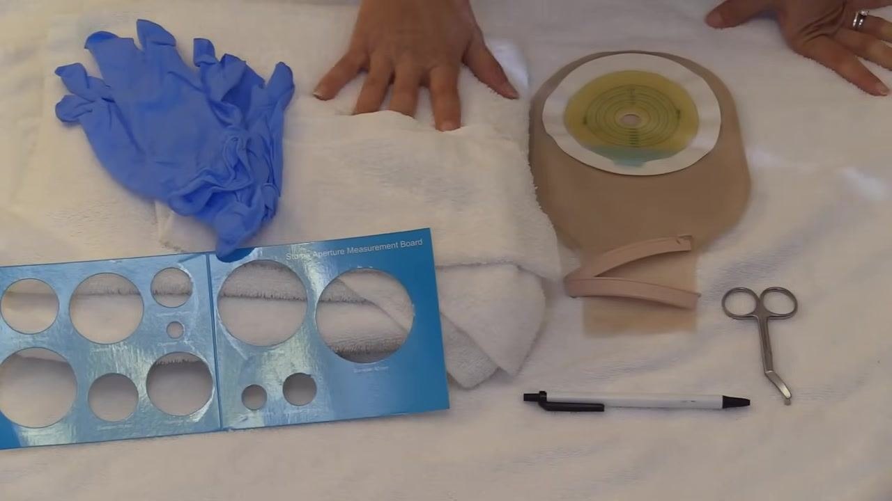 Gloved hands holding a full ostomy pouch attached to a stoma model, showing a pouch that needs to be emptied or changed