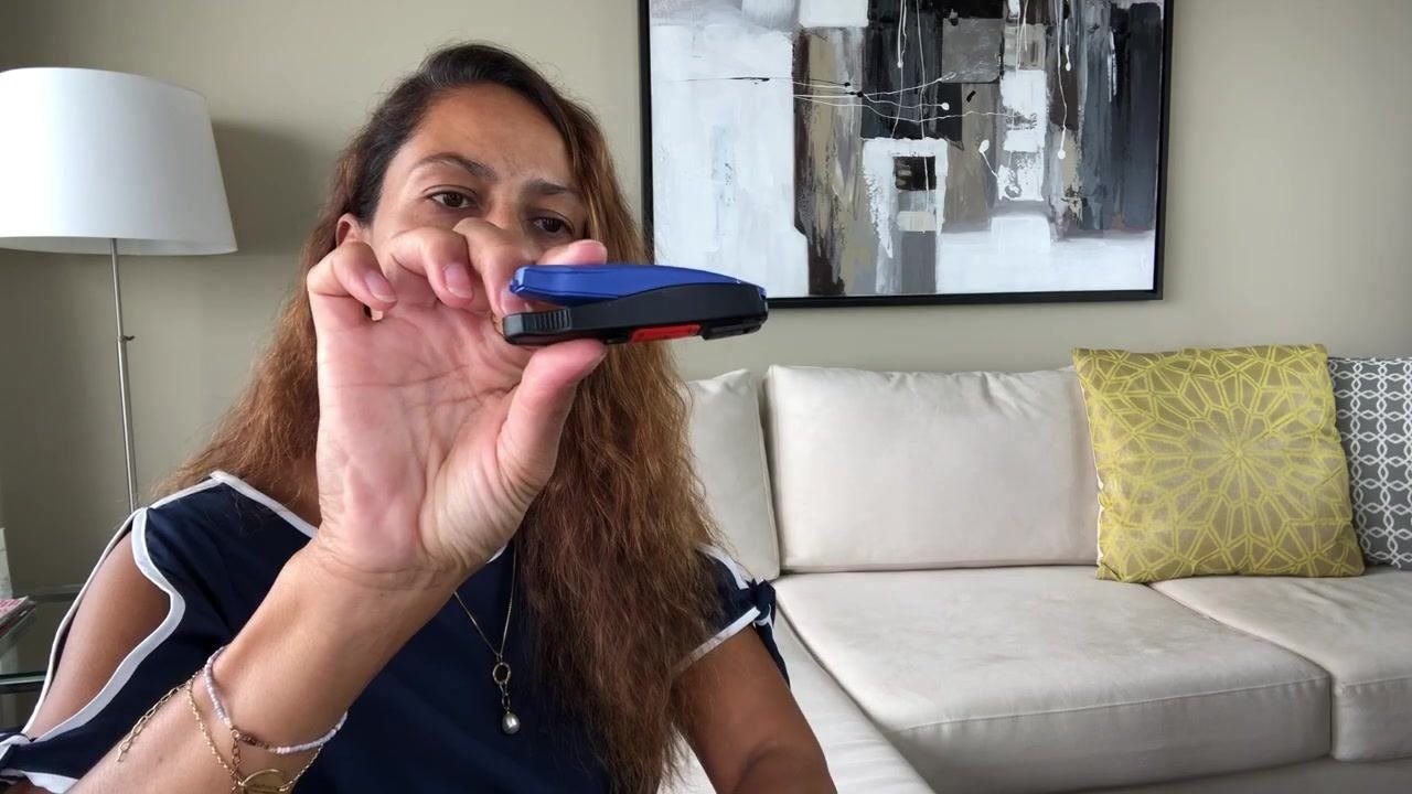 Person holding a blue BD Needle Clipper and demonstrating how to open it by pressing the sides. The device is shown in a living room setting with a couch and pillows in the background.