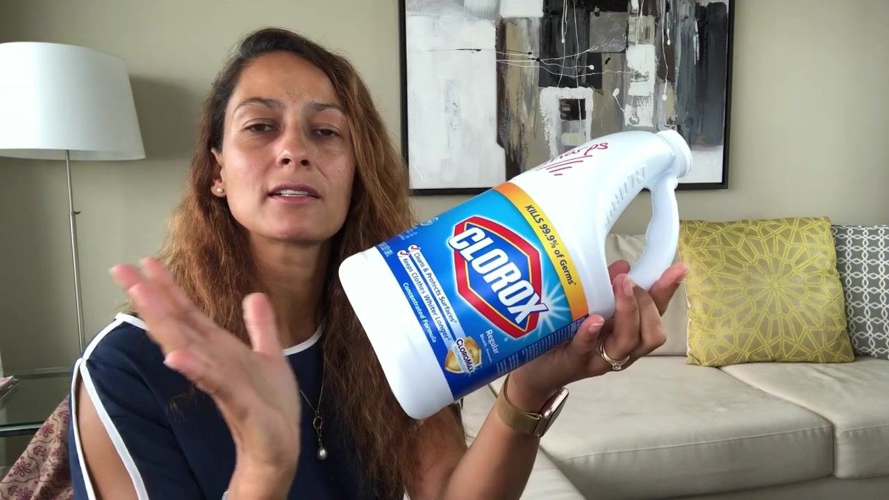 Person holding a clean, empty Clorox bleach bottle labeled "Sharps" in red marker, shown as an example of a homemade sharps container, in a living room setting with a couch, pillows, and a lamp in the background.